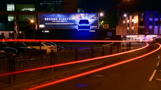 A highway lit with electrical lights and has a electrically glowing creative billboard advertising of BMW i3